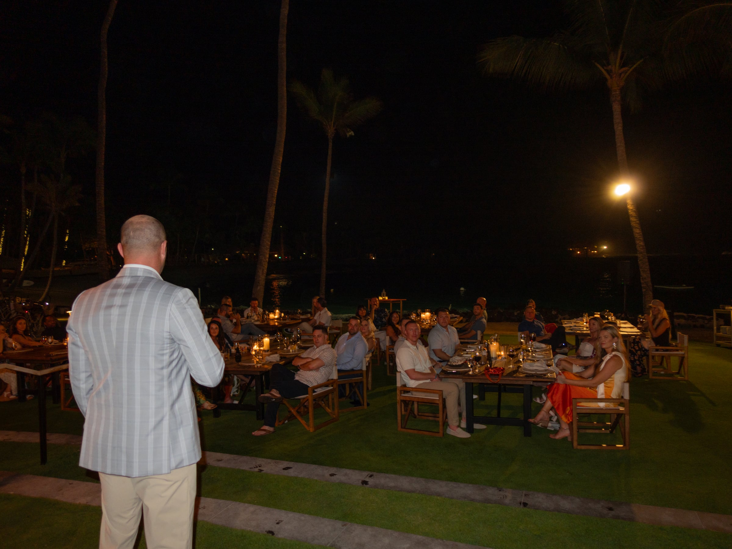 Guests seated at candlelit tables on the lawn for the opening dinner, palms and the bay beyond